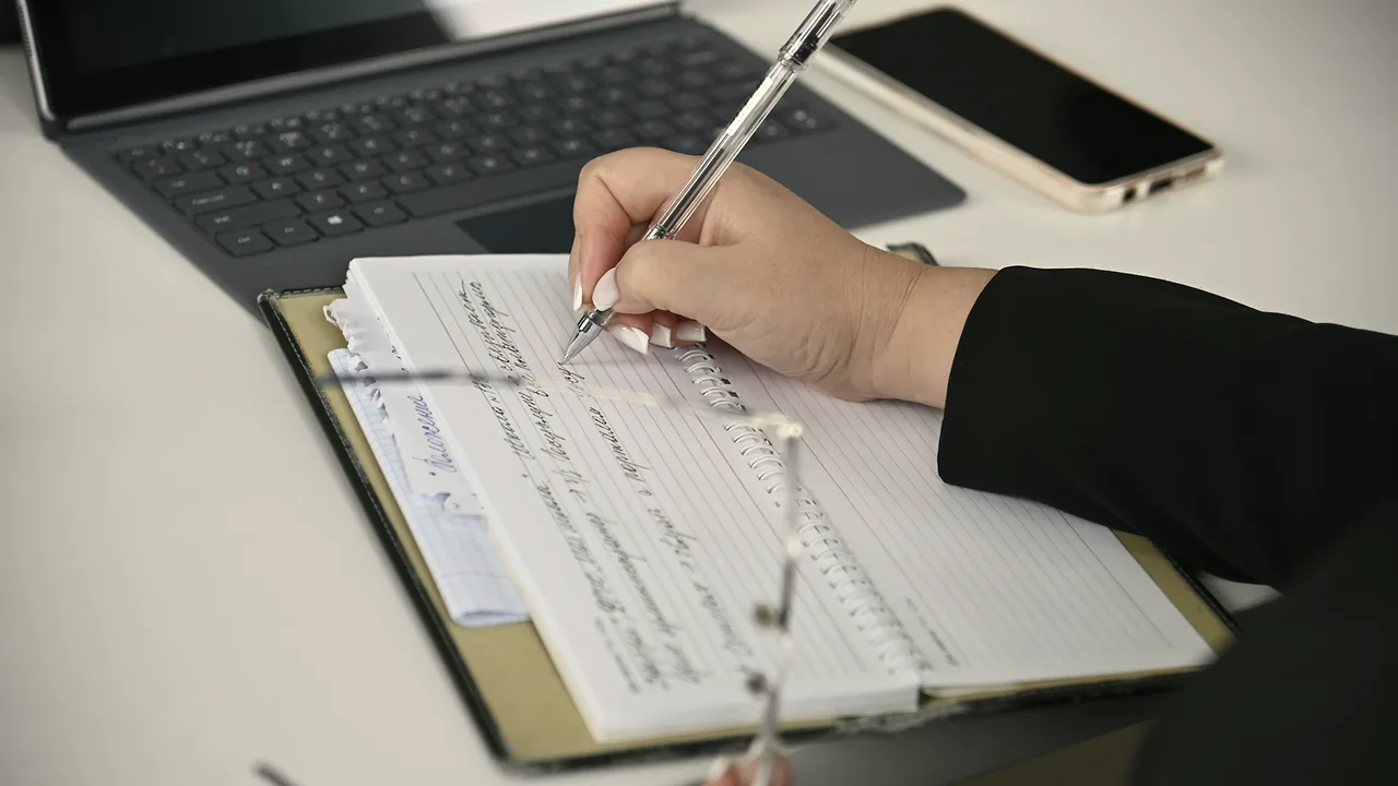 A hand writing notes in a ring-bound notebook in front of a laptop and a phone