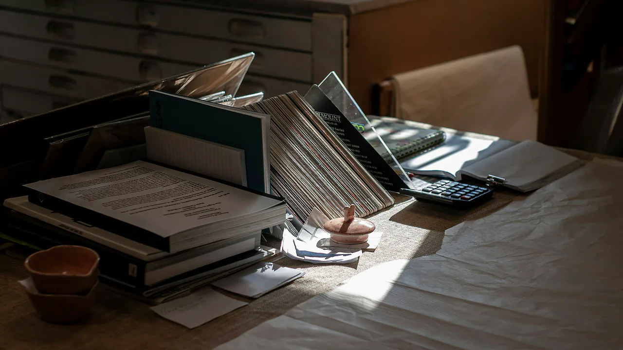 Afternoon sun across a desk stacked with papers, files in a rack and a calculator